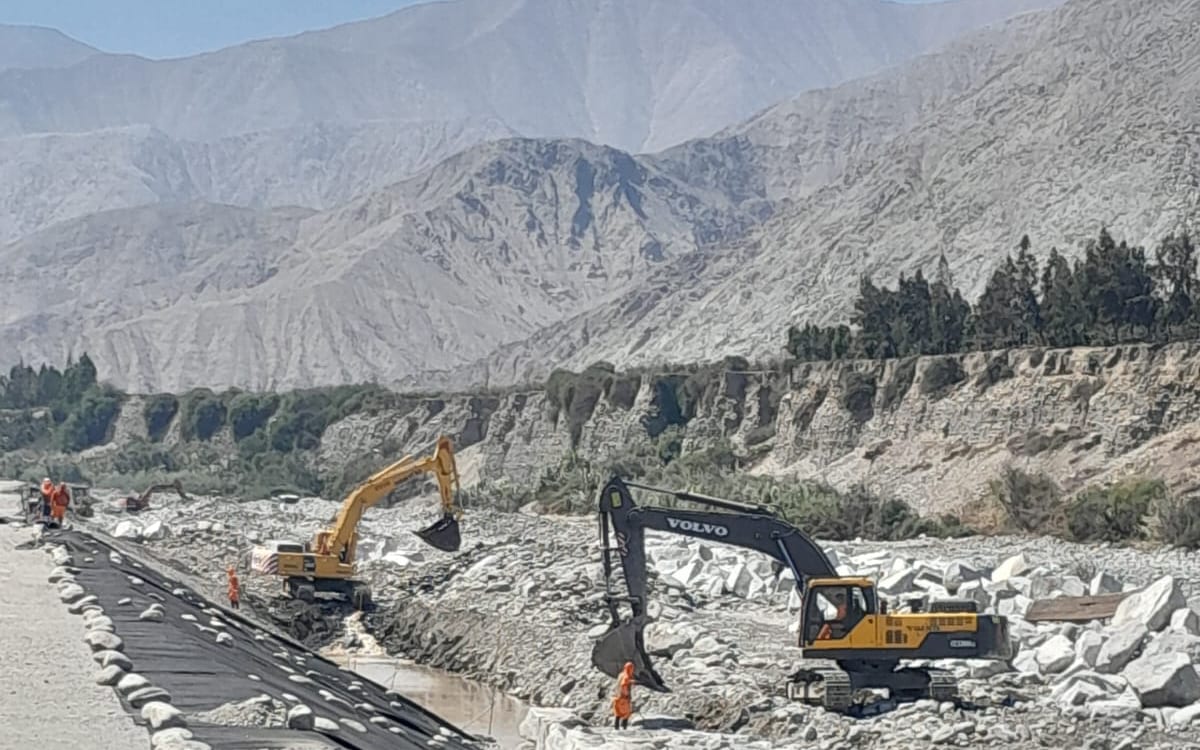 Dos excavadoras y personal trabajando en un cauce, con montañas al fondo.